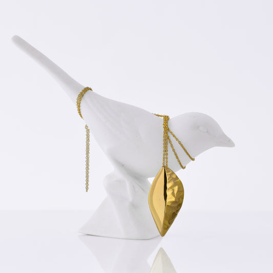 An 18kt Fairmined yellow gold leaf-shaped pendant on an 18" Fairmined 18kt yellow gold chain against a white background, draped over a porcelain white bird.